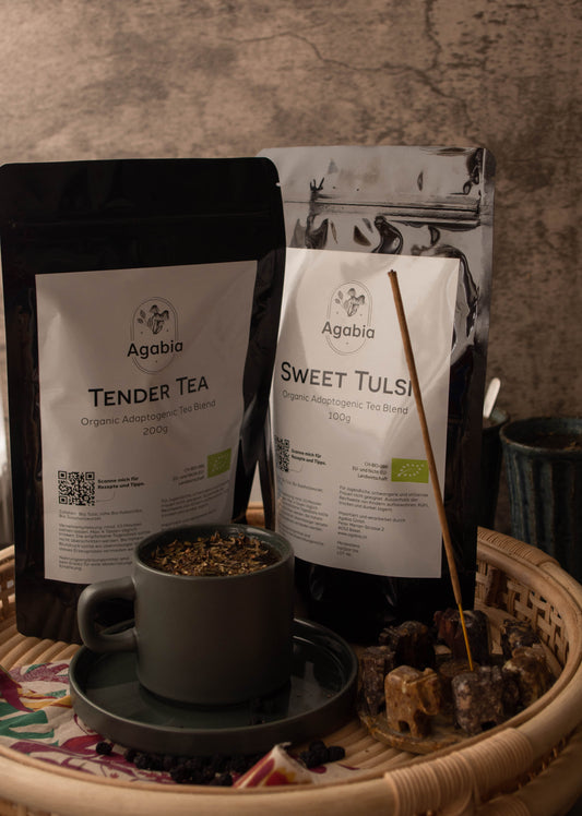 Ein Päckchen "Tender Tea" und ein Päckchen "Sweet Tulsi" auf einem Rattantablett. Vor den Teepaketen steht eine graue Keramiktasse auf einer Untertasse, gefüllt mit Tulsi-Tee. Neben der Teetasse brennt Weihrauch in einem Brenner in Form einer Reihe von Elefanten.